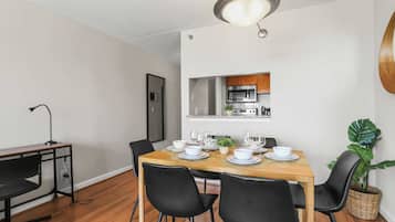Comedor con una mesa de madera dispuesta para seis comensales, sillas negras, una lámpara colgante y una cocina visible a través de una puerta abierta.