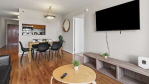 Ett modernt vardagsrum med en platt-TV, en matplats med ett träbord och stolar, ett kök med mikrovågsugn och skåp samt en hall med en dörr.