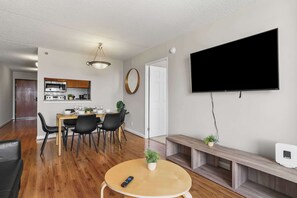 Een moderne woonkamer met een flatscreen-tv, een eethoek met een houten tafel en stoelen, een keuken met een magnetron en kasten, en een gang met een deur.