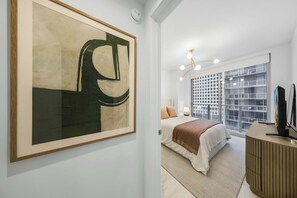Ein modernes Schlafzimmer mit einem großen abstrakten Gemälde, einem Bett mit einer beige-braunen Streifendecke, einem Nachttisch mit Lampe und einem Holzschrank mit Fernseher.