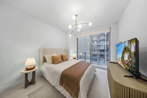 Ein modernes Schlafzimmer mit einem Bett, Nachttischlampen, einem Fernseher und einem großen Fenster mit Blick auf Gebäude.