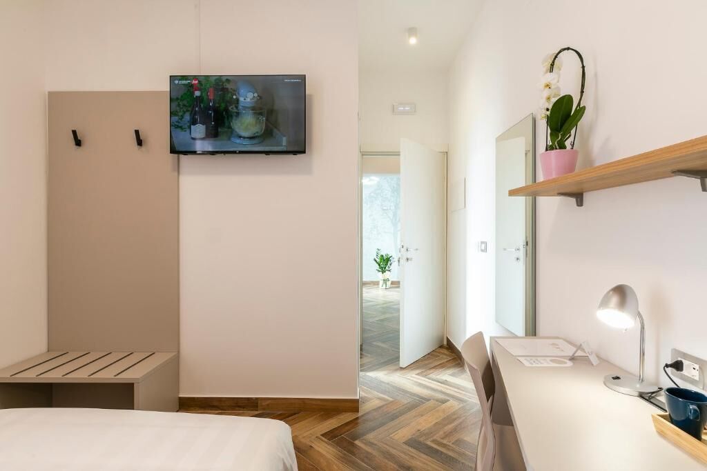 Et moderne hotelværelse med en seng, et skrivebord, et tv ophængt på væggen, en potteplante og en træhylde.