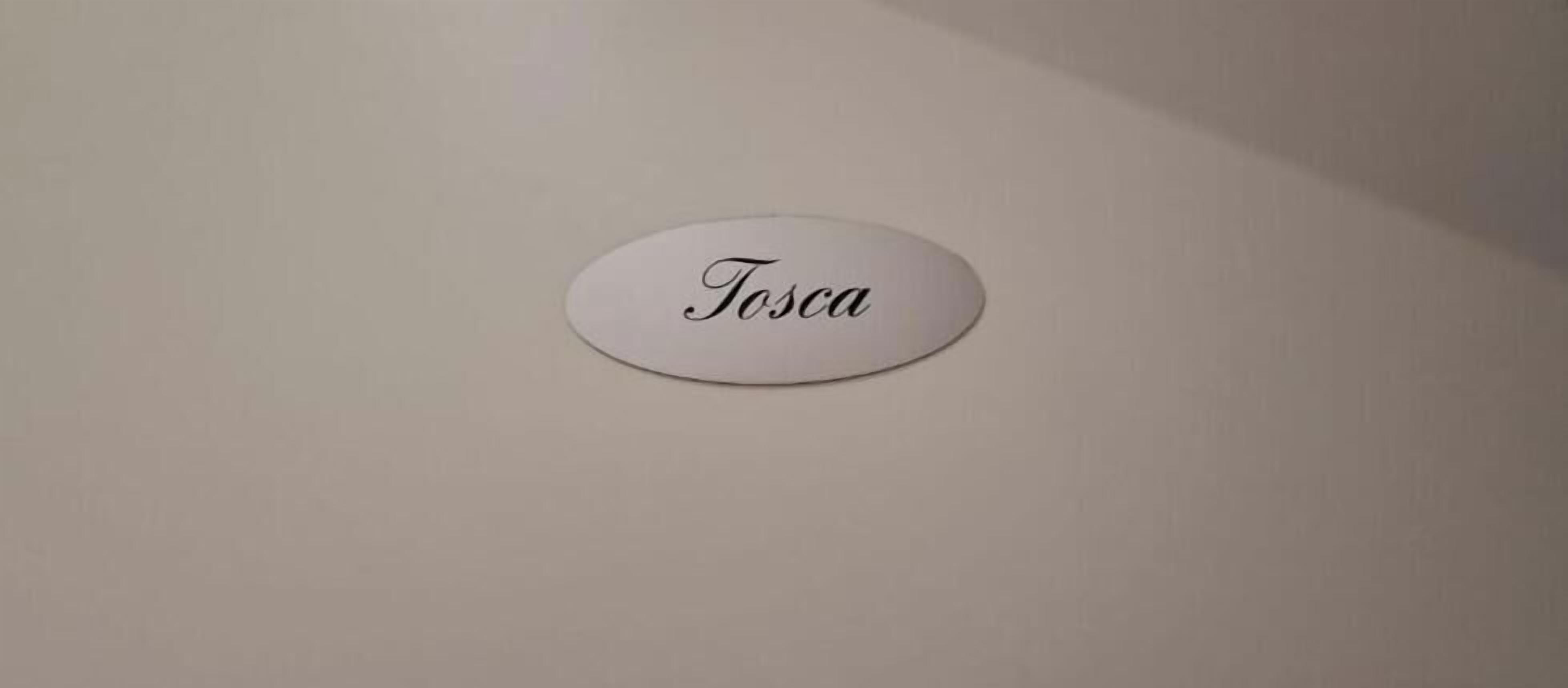 Una señal blanca y ovalada con el nombre 'Tosca' sobre una pared lisa.