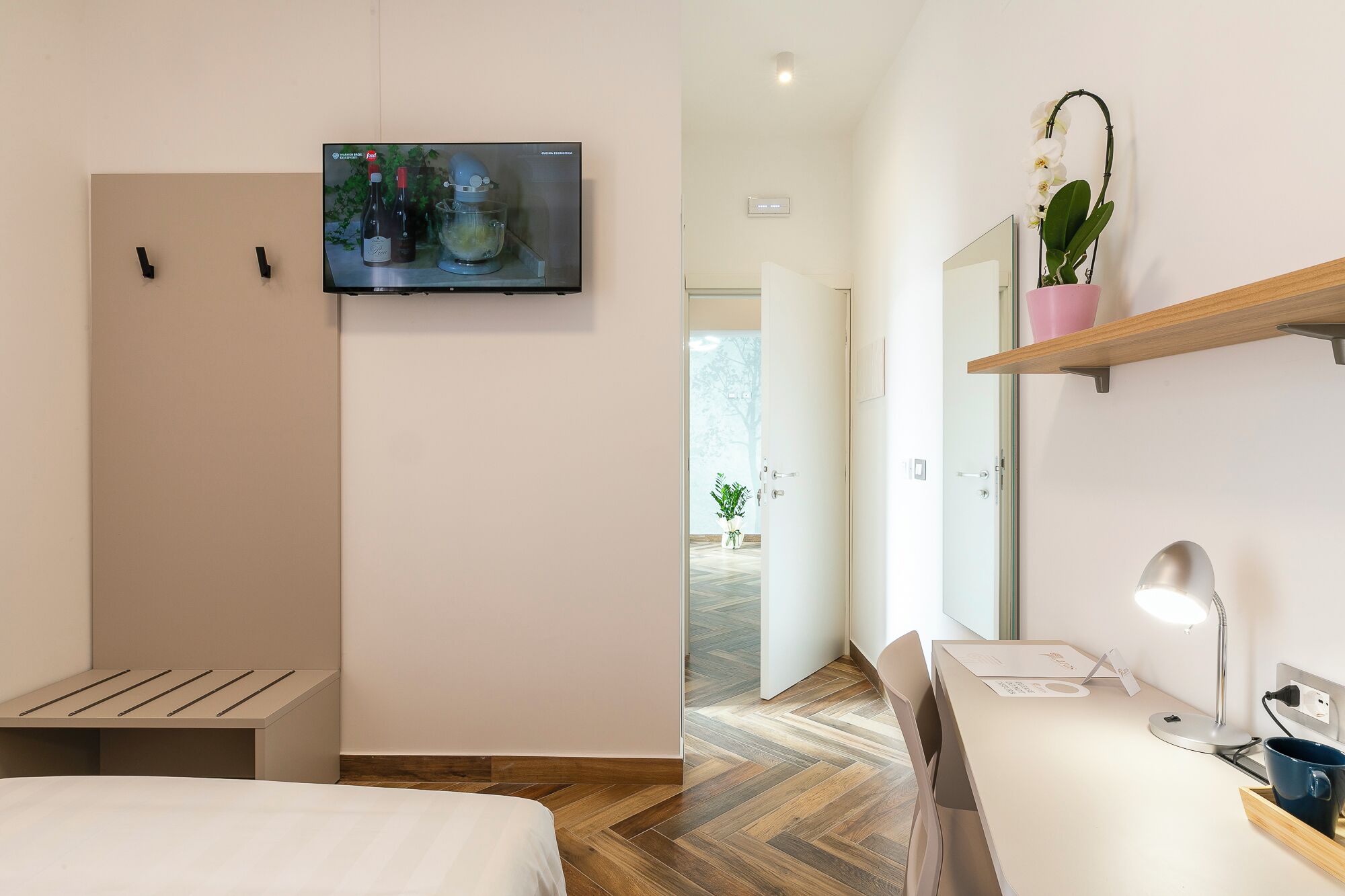 Une chambre d’hôtel avec un lit, un téléviseur fixé au mur, un bureau avec une lampe et une plante en pot.