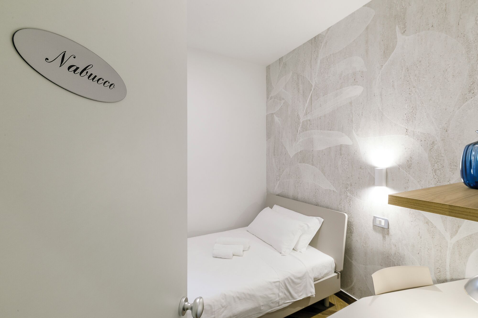 A hotel room with a bed, a wall-mounted light, a wooden shelf, and a sign with the name 'Nabucco'.