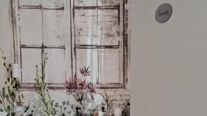 Una habitación con piso de espiga, una puerta blanca con un letrero, una ventana con aspecto desgastado y una pequeña mesa con una silla.