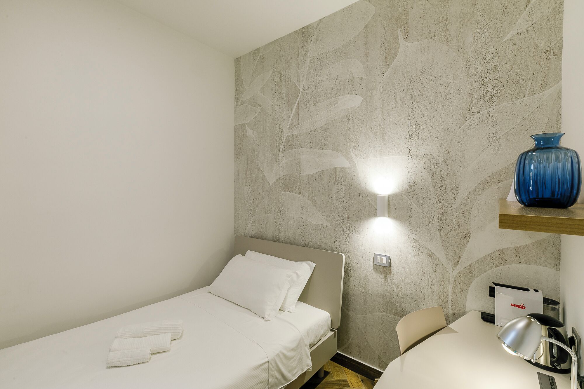 A small, single-bed room with a textured wall, a bedside table with a lamp, and a blue vase on a shelf.