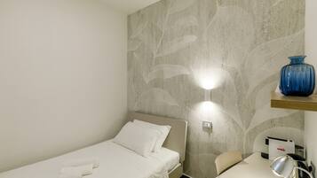 A small, single-bed room with a textured wall, a bedside table with a lamp, and a blue vase on a shelf.