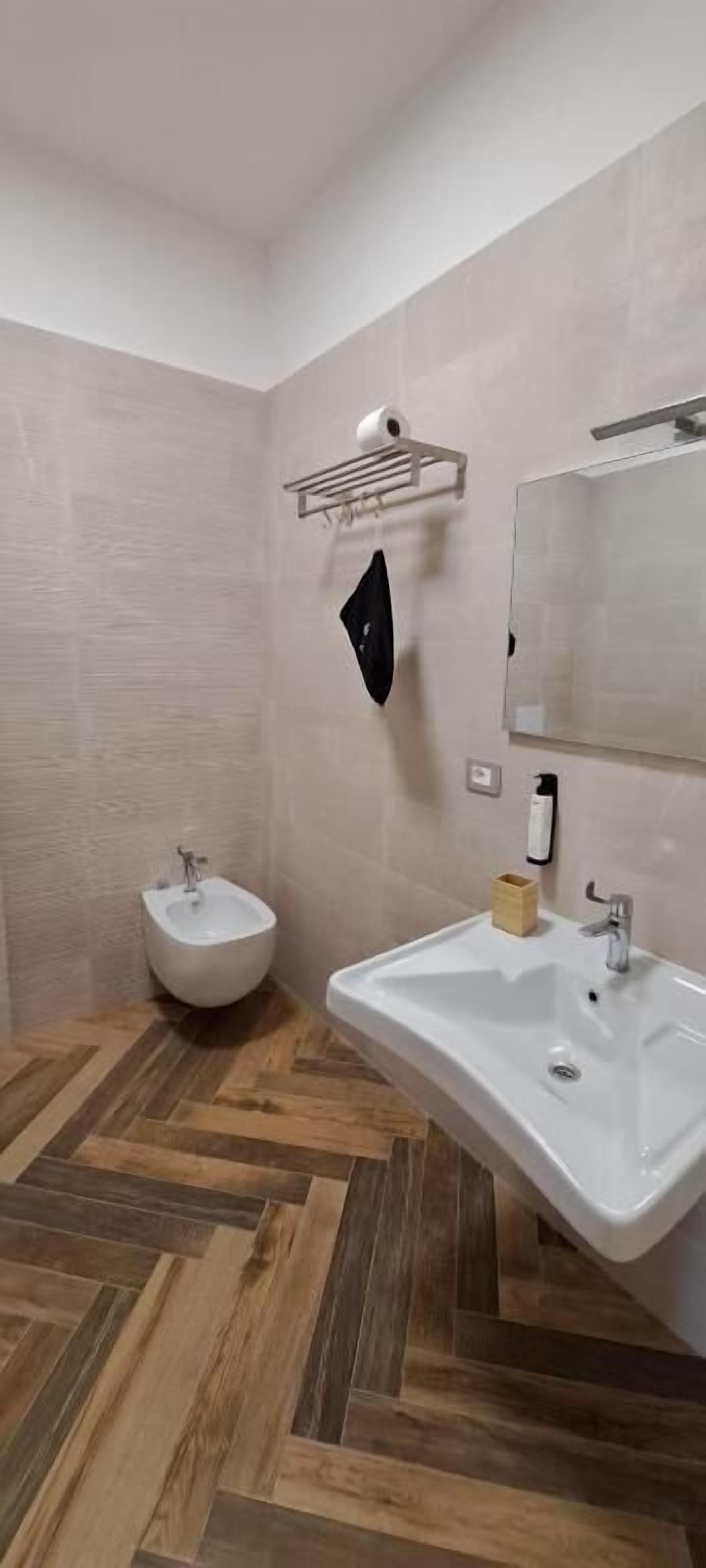 Un baño con piso de madera en patrón de espiga, un bidet blanco, un lavamanos blanco con dispensador de jabón y un perchero para toallas.