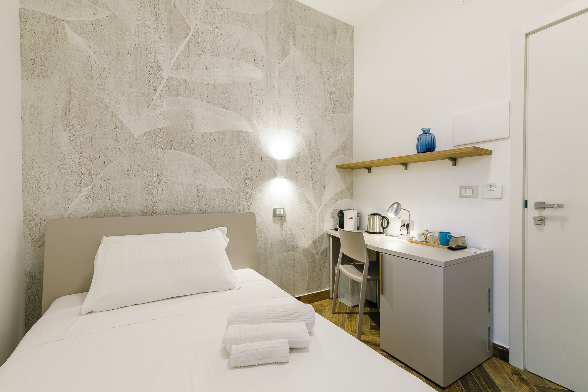 A hotel room with a bed, a desk, a chair, a small kitchenette, and a wardrobe.