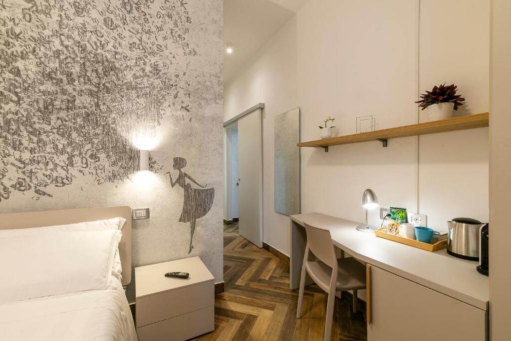 A hotel room with a bed, a desk with a chair, a small table with a kettle, a shelf with a plant, and a wall with a mural.