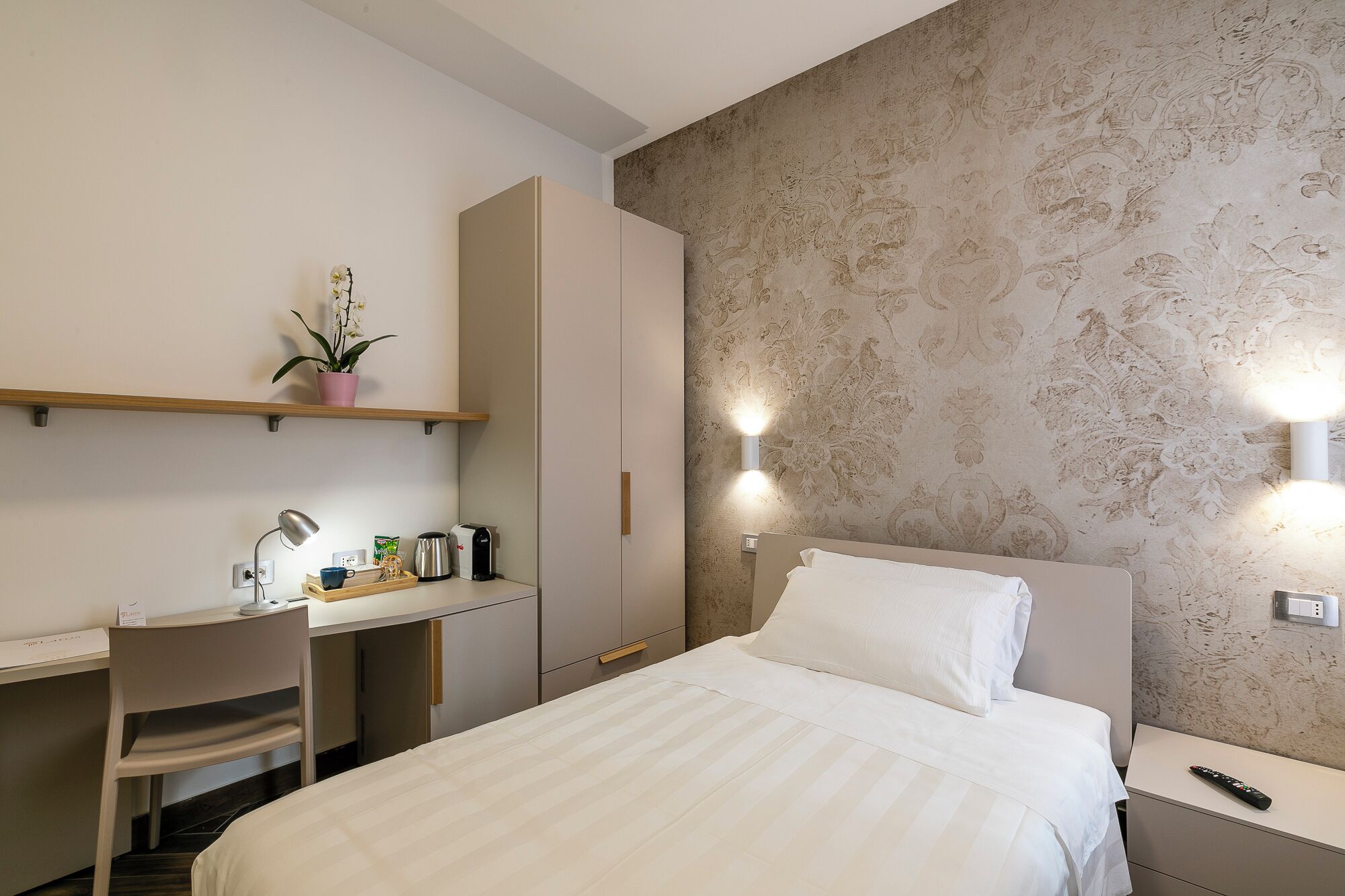 Ein Hotelzimmer mit einem Bett, einem Schreibtisch, einem Stuhl, einem Regal mit einer Pflanze und einer Wandleuchte.