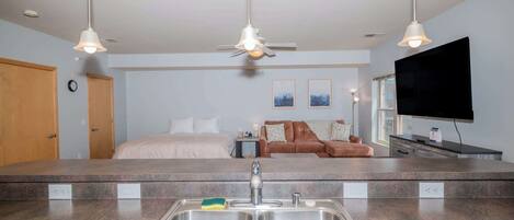 A modern hotel room with a kitchenette, a bed, a seating area, and a flat-screen TV.
