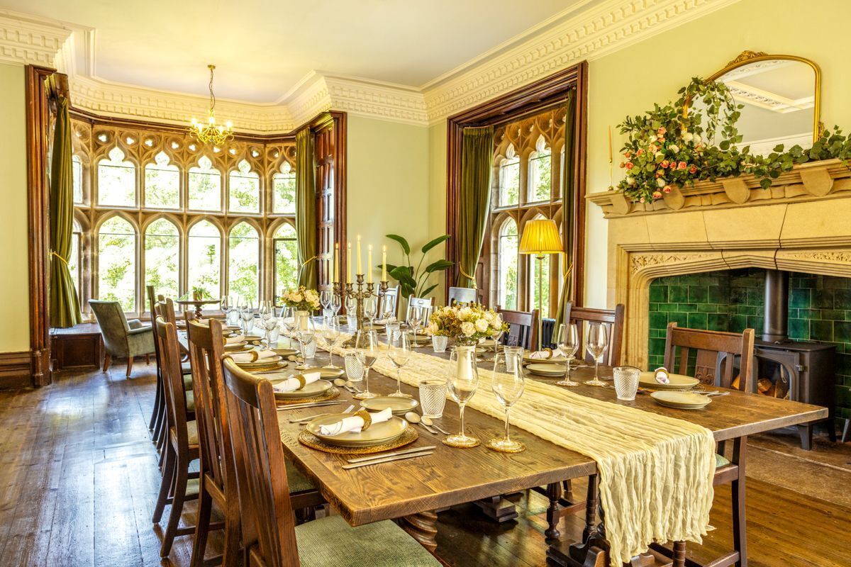 Une salle à manger avec une longue table en bois dressée pour le repas, de grandes fenêtres et une cheminée.