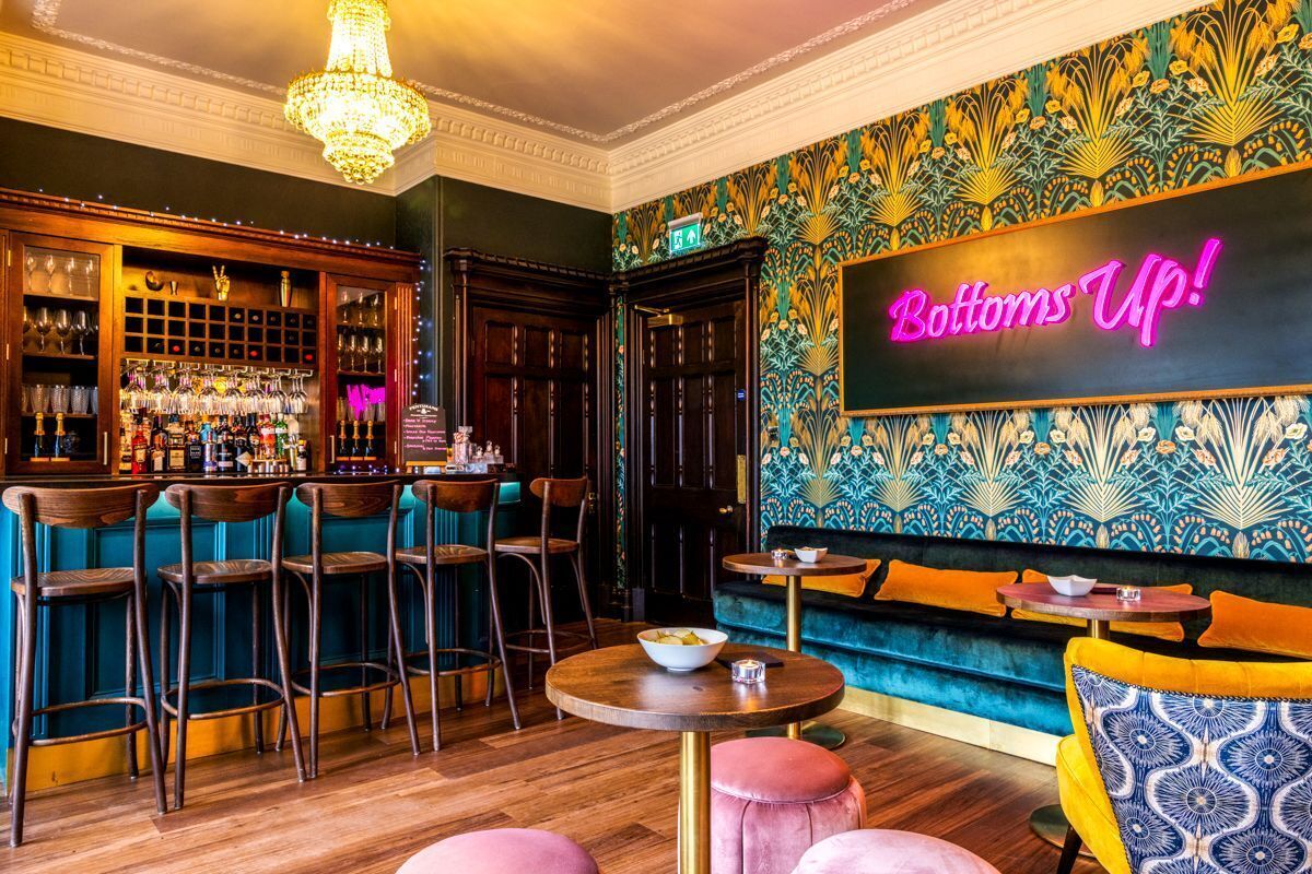 A bar area with stools, a bar counter, and a neon sign that reads 'Bottoms Up!'.