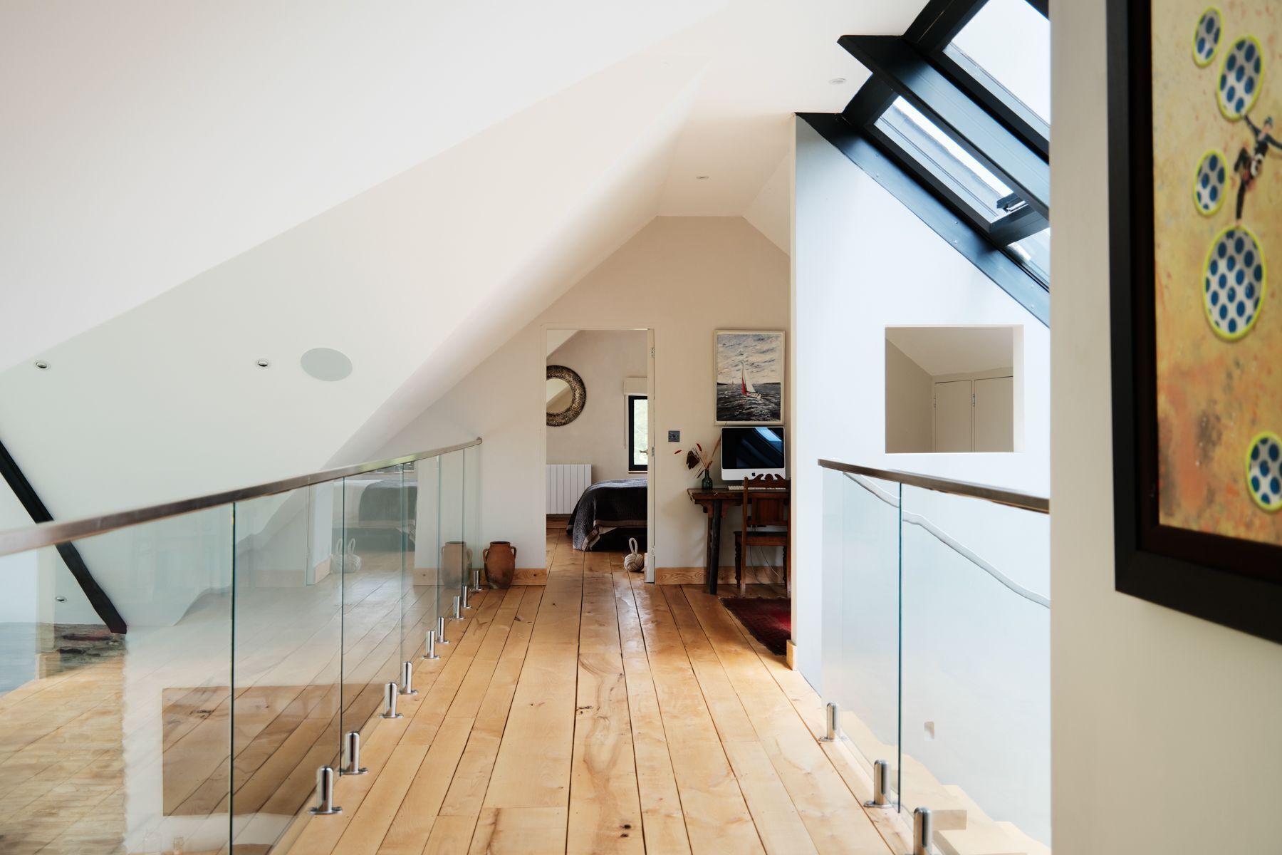 A modern interior with a wooden floor, glass railing, and a view of a bedroom with a bed and a painting.