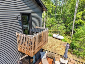 Une terrasse en bois avec une table et des chaises, une petite table d’extérieur et une bouteille de gaz marquée « LAKES GAS », le tout situé dans une zone boisée.