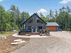 A modern two-story house with a large deck, a garage, and a forested backdrop.