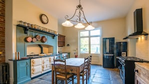 Une cuisine avec un sol en pierre, une table à manger avec des chaises, un fourneau, un réfrigérateur et une fenêtre donnant sur l’extérieur.