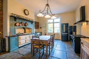 A kitchen with a stone floor, a dining table with chairs, a stove, a refrigerator, and a window with a view of the outdoors.