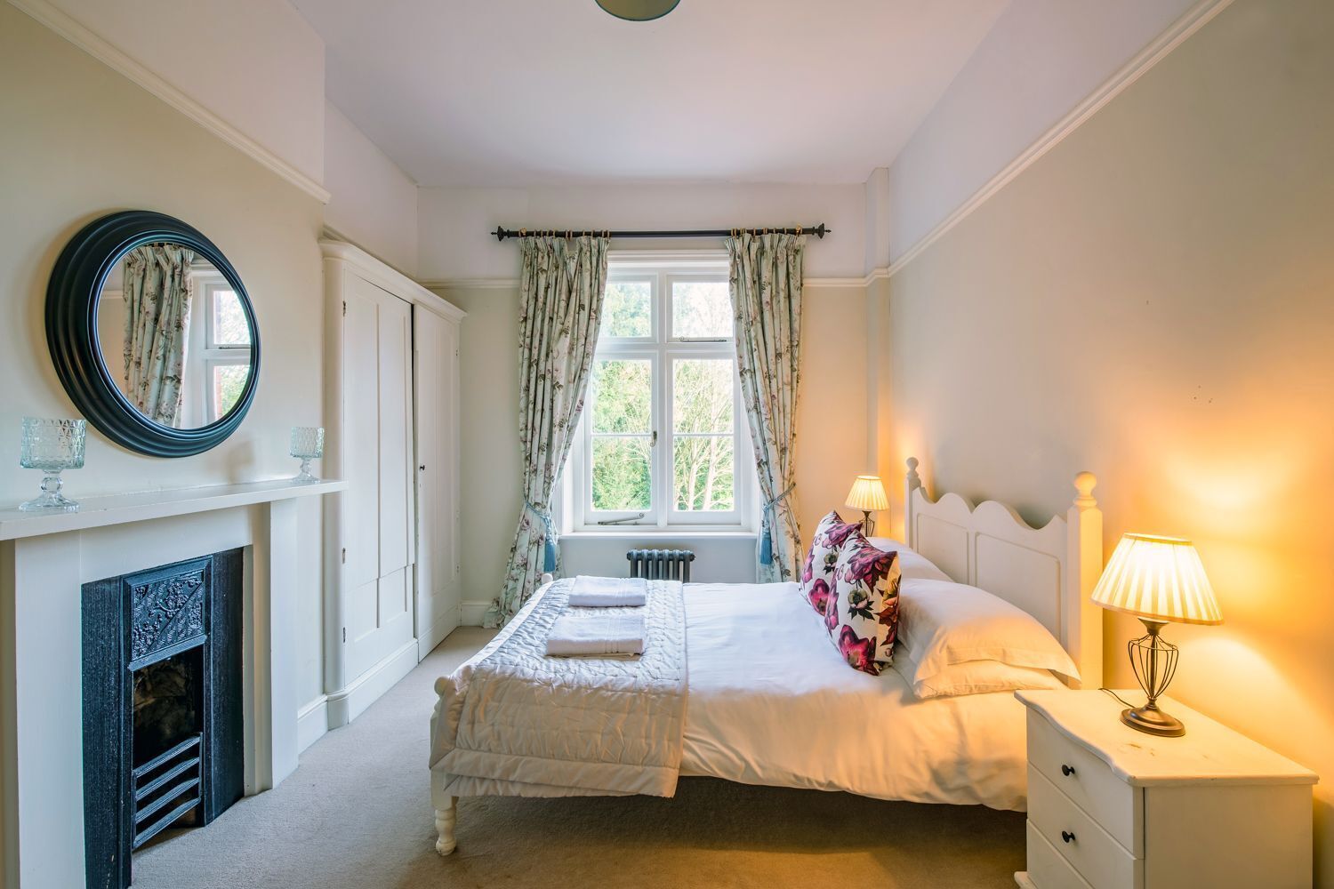 A bedroom with a bed, a nightstand, a lamp, a mirror, a fireplace, and a window with curtains.