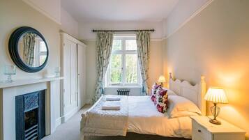 A bedroom with a bed, a nightstand, a lamp, a mirror, a fireplace, and a window with curtains.