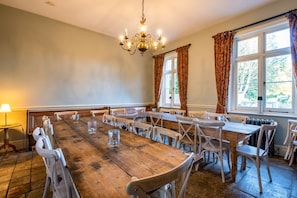 A dining room with a large wooden table, white chairs, a chandelier, and a view through a window.