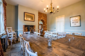 A dining room with a large wooden table, white chairs, a brick fireplace, and a chandelier.