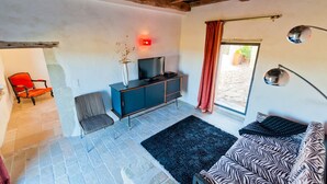 A living room with a stone floor, a blue sofa, a red chair, a television on a cabinet, and a window with a red curtain. - Mounicat - Four Bedroom Villa, Sleeps 10 (Baleyssagues)