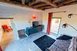 A living room with a stone floor, a blue sofa, a red chair, a television on a cabinet, and a window with a red curtain. - Mounicat - Four Bedroom Villa, Sleeps 10 (Baleyssagues)