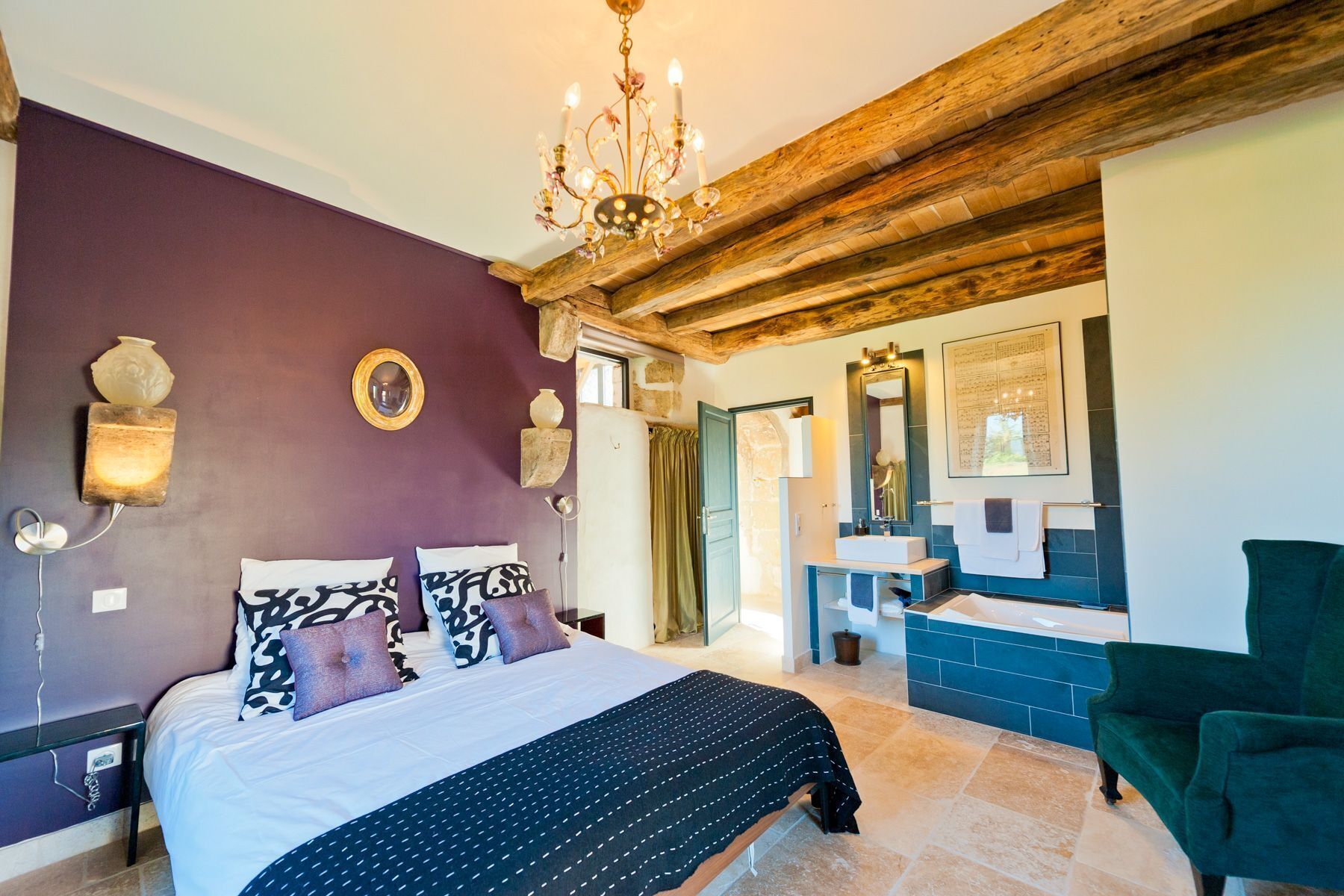 Une chambre à coucher avec un lit, un lustre, une salle de bain avec un lavabo et un miroir, et un fauteuil vert.