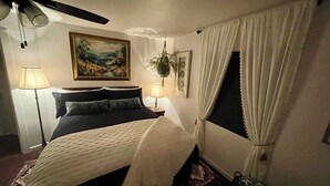 A bedroom with a bed, a ceiling fan, a painting, a plant, and a window with curtains. - Two Sisters Inn (Ahwahnee)