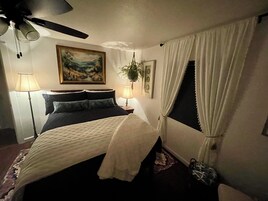 Un dormitorio con cama, ventilador de techo, un cuadro, una planta y una ventana con cortinas.