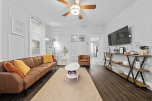 Ein modernes Wohnzimmer mit einem braunen Ledersofa, einem weißen Couchtisch, einem Deckenventilator und einem an der Wand montierten Fernseher.