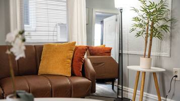 A brown leather sofa with orange and yellow cushions, a white round table, and a potted plant on a white pedestal.