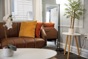 A brown leather sofa with orange and yellow cushions, a white round table, and a potted plant on a white pedestal. - Two Bedrooms • Coffee Station • Family Friendly (Jersey City)