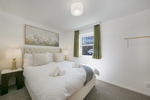 A hotel room with a bed, a nightstand, a lamp, a painting, a window with curtains, and a view of the street. - Chapel Street Suite - Grampian Lettings (Aberdeen)