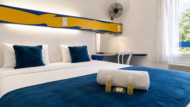 Une chambre d’hôtel comprenant un lit, un bureau, une chaise, une fenêtre avec des rideaux et un ventilateur fixé au mur.