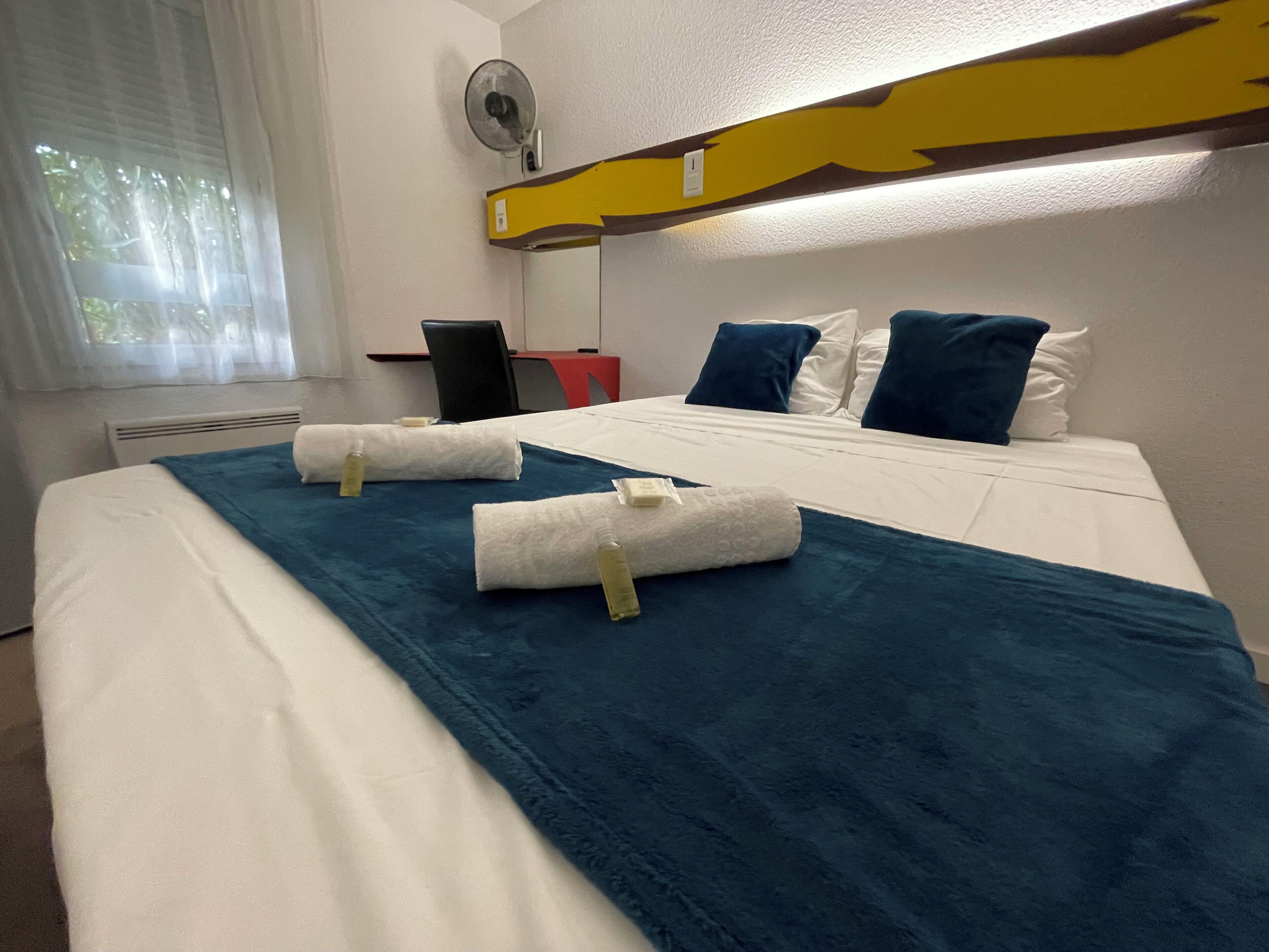 A hotel room with a bed, a desk, a chair, a window with curtains, and a wall-mounted fan.