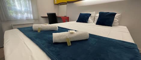 A hotel room with a bed, a desk, a chair, a window with curtains, and a wall-mounted fan.
