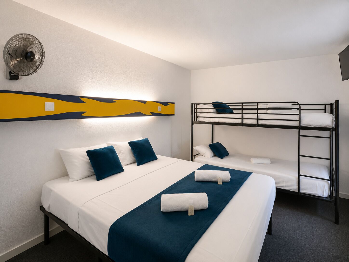 A hotel room with a bunk bed, a single bed, and a wall-mounted fan.