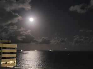 Nachtszene mit Vollmond, einem Gewässer und einem Gebäude mit beleuchteten Fenstern.