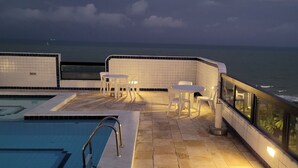 Un área de piscina en la azotea con vistas al océano, que incluye una piscina, sillas de descanso y una pequeña mesa.