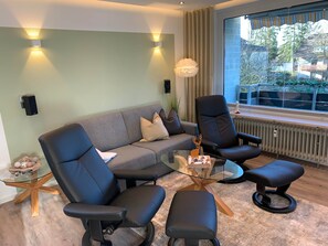 Ein modernes Wohnzimmer mit einer Couch, zwei Sesseln, einem Glastisch und Blick ins Freie.