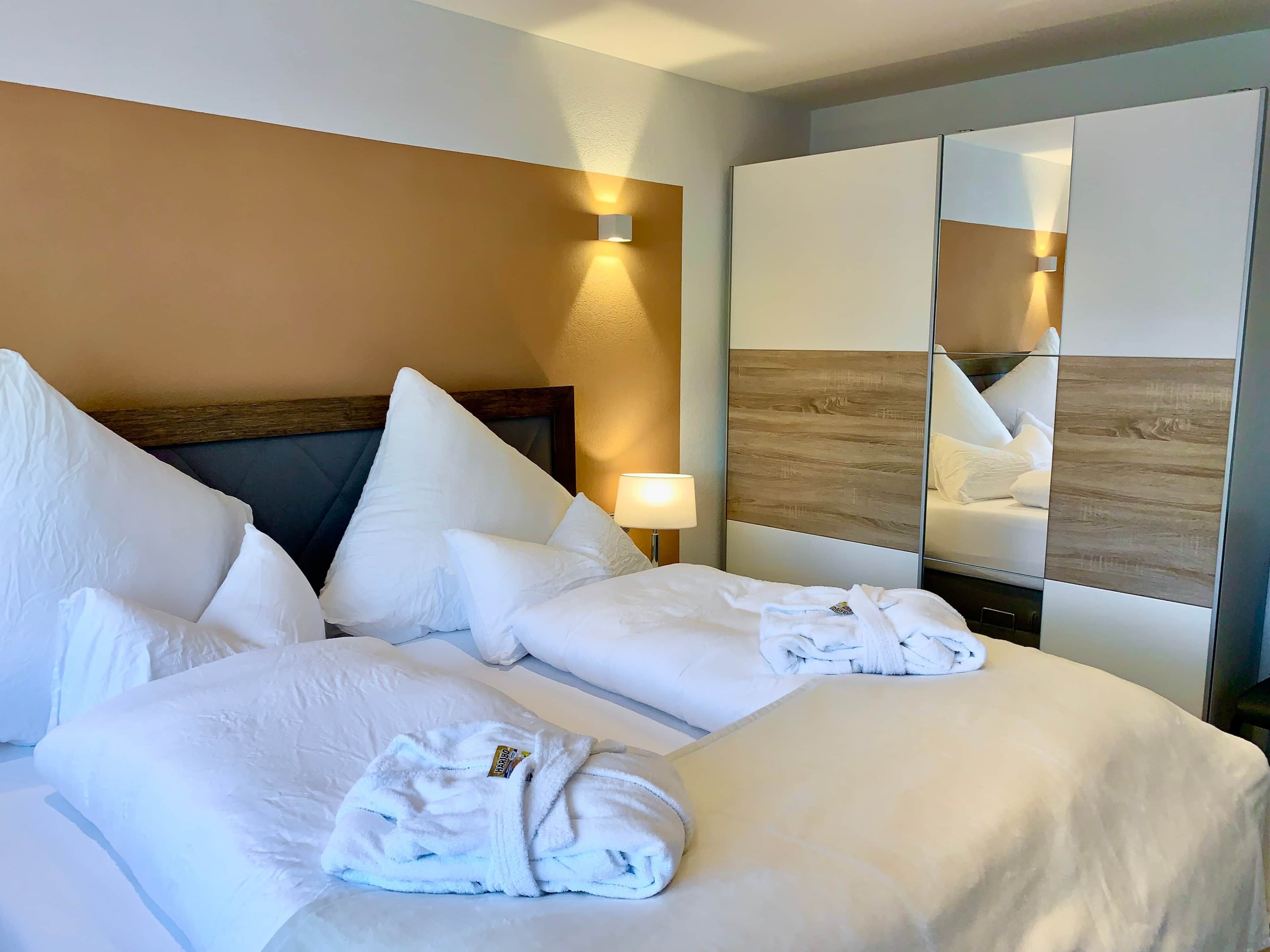 Une chambre d’hôtel avec un grand lit, plusieurs oreillers, une lampe de chevet, un miroir et un peignoir posé sur le lit.