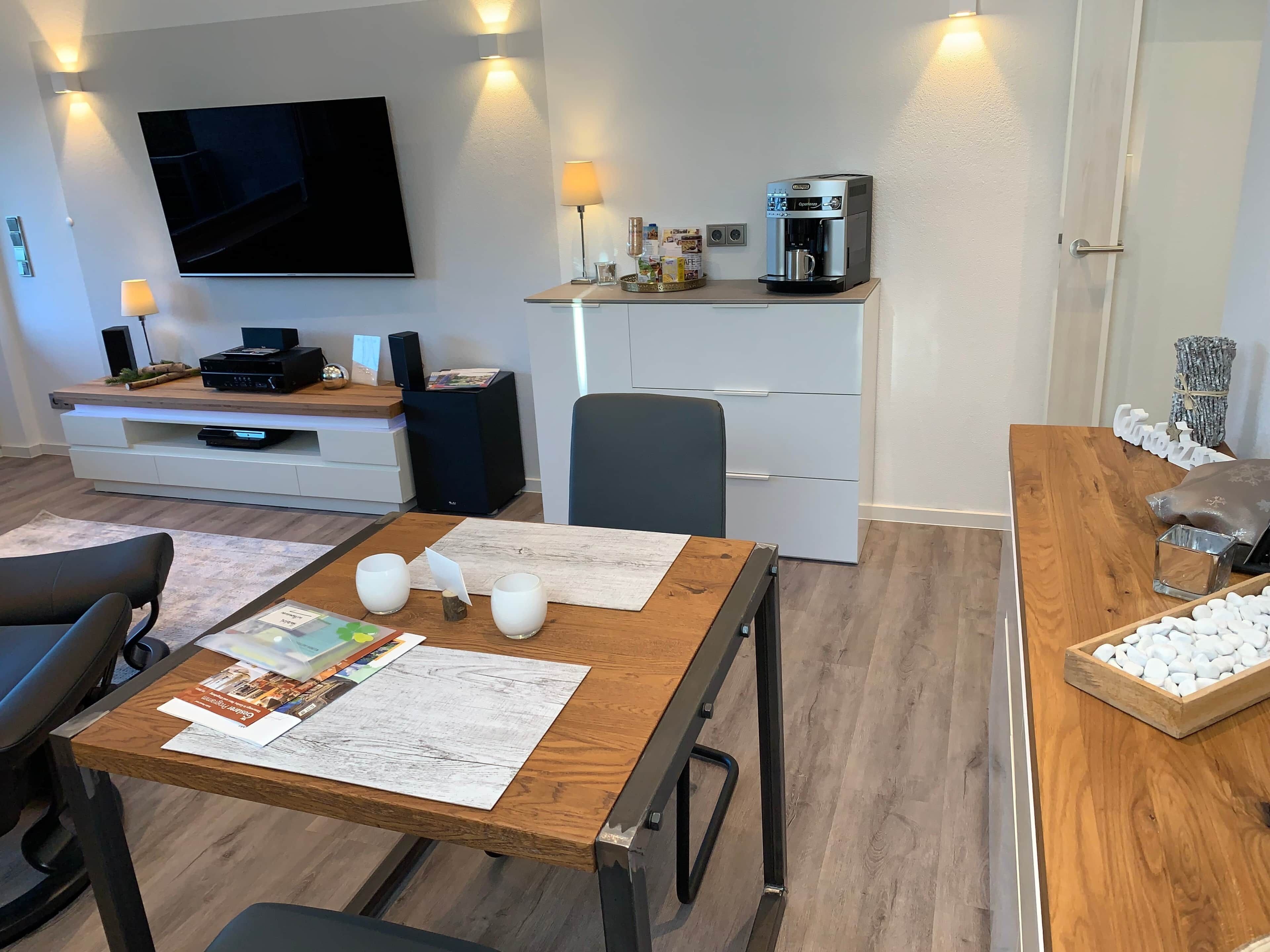 A modern living room with a flat-screen TV, a coffee machine, and a dining area with a wooden table and chairs.