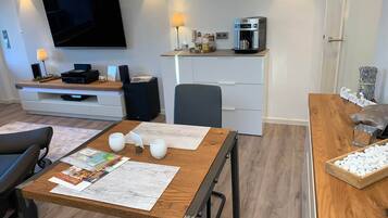 A modern living room with a flat-screen TV, a coffee machine, and a dining area with a wooden table and chairs.