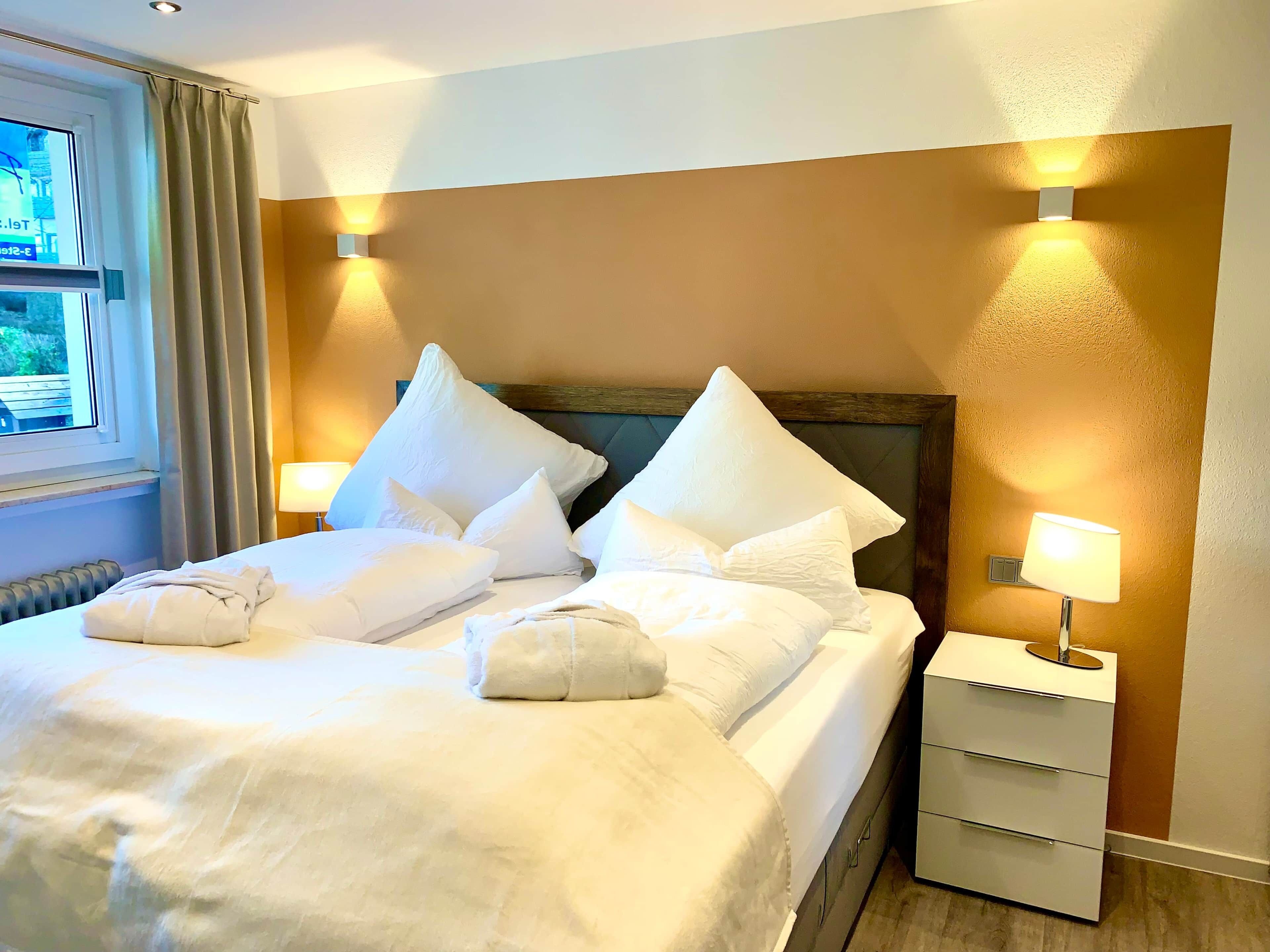A hotel room with a bed, pillows, a nightstand, and two wall-mounted lamps.