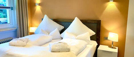 Quarto de hotel com cama, almofadas, mesa de cabeceira e duas luminárias fixadas na parede.