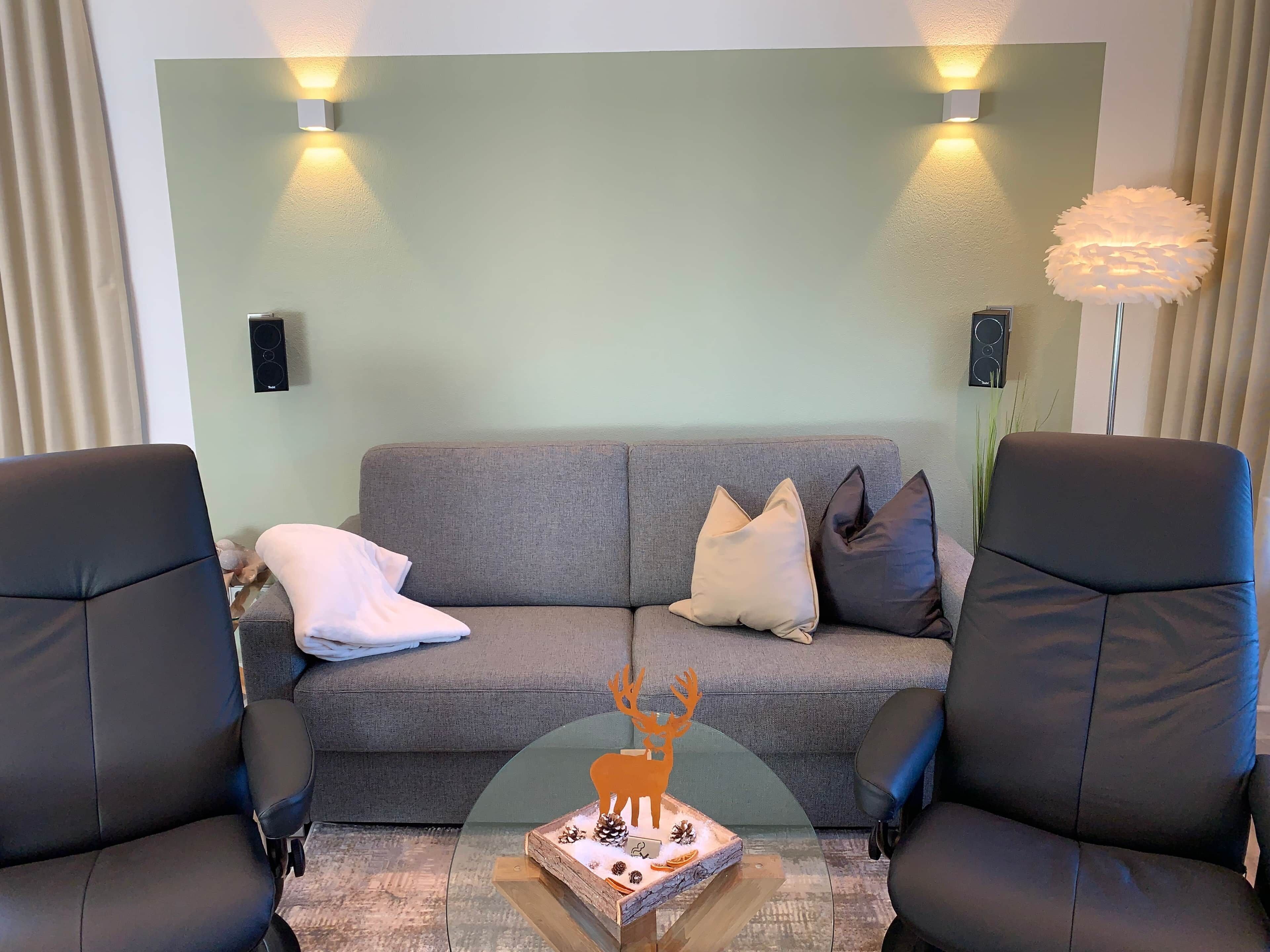 Ein Wohnzimmer mit grauem Sofa, zwei schwarzen Sessel, einem Couchtisch mit Dekoration und Wandleuchten.